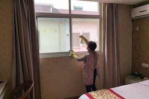 Una persona limpiando un ventanal en una habitación con cama, sillón y aire acondicionado.