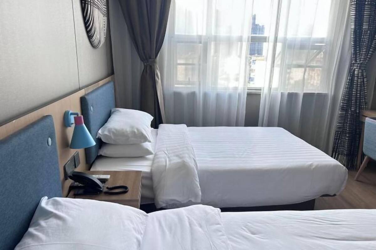 A hotel room with two beds, a desk with a lamp, a phone, and a view of the city through large windows.