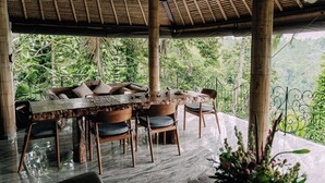 Un espace extérieur abrité, aménagé pour les repas, avec une table et des chaises en bois, entouré d’une végétation luxuriante et délimité par une rambarde en métal.