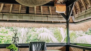 Une pièce avec un bureau en bois, un fauteuil de bureau noir, un luminaire suspendu en fibres tressées et une vue sur des palmiers par la fenêtre.