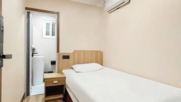Een kleine kamer met een enkel bed, een houten hoofdbord, een nachtkastje, een lade en een witte, aan de muur bevestigde airconditioner.
