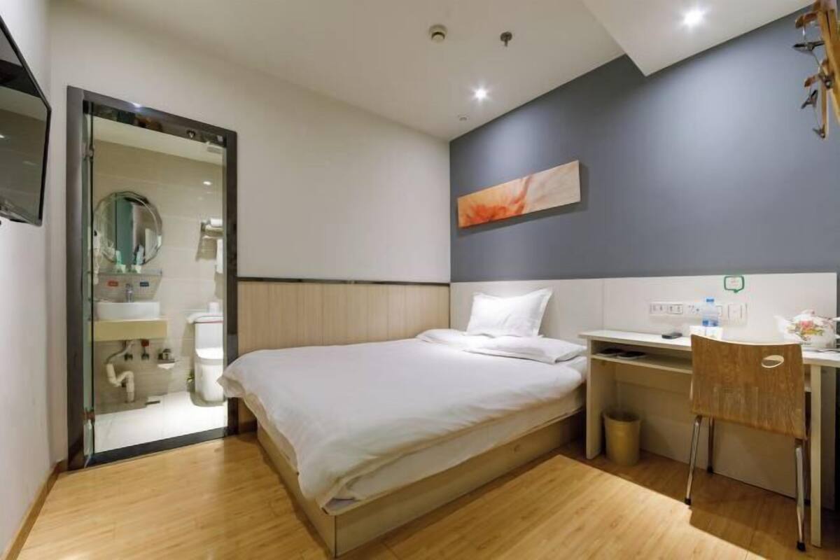 A hotel room with a bed, a desk, a chair, a mirror, and a shower area.