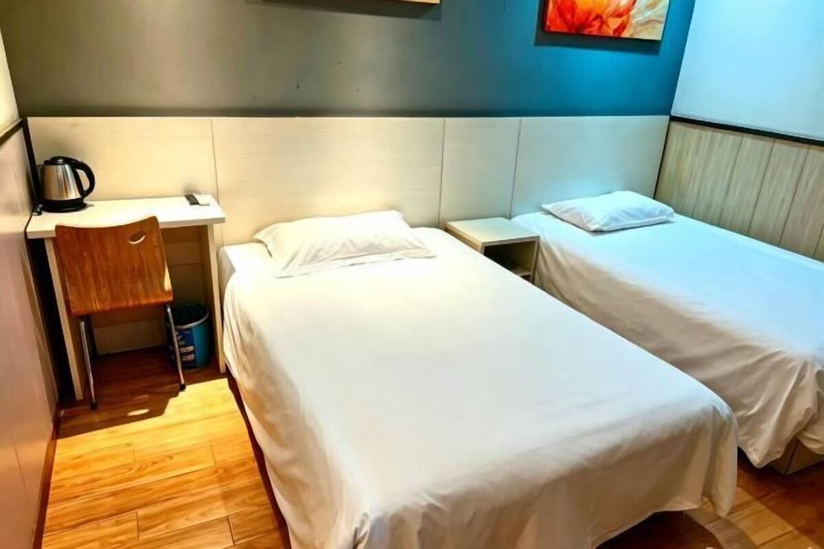 A hotel room with two single beds, a small desk with a kettle, and a chair.