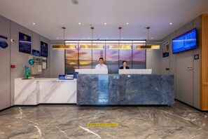 Zona de recepción de un hotel con un mostrador de mármol, dos recepcionistas, una pantalla de televisión grande y una pared con carteles informativos.