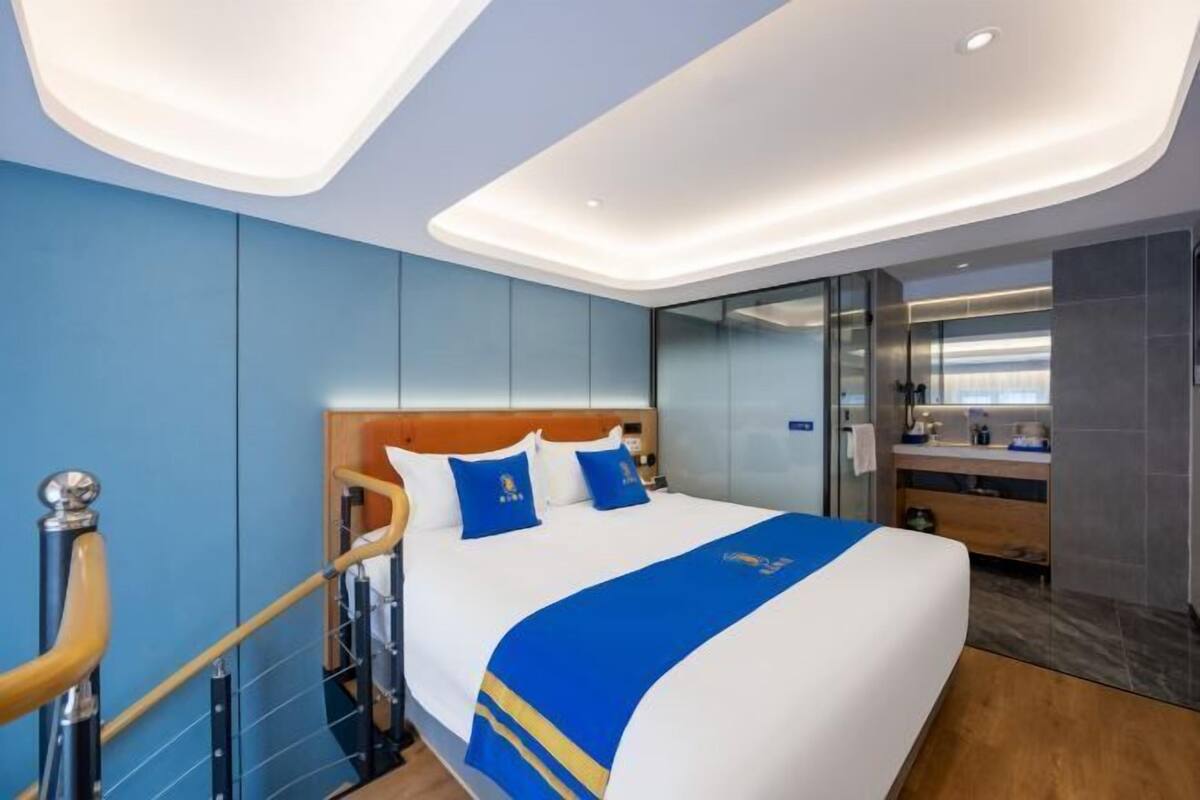 A modern hotel room with a large bed, a wooden headboard, a glass shower enclosure, and a wooden staircase.