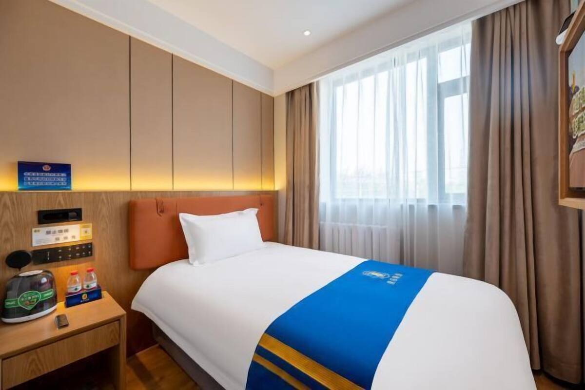 A hotel room with a bed, a nightstand with a coffee maker, a control panel, and a window with curtains.