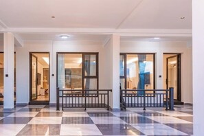 Un hall d’hôtel avec un sol à carreaux, un banc central et des portes vitrées donnant accès aux chambres.
