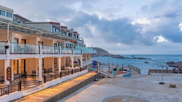 Un hotel frente al mar con varios balcones y una zona exterior cubierta, además de un camino pavimentado.