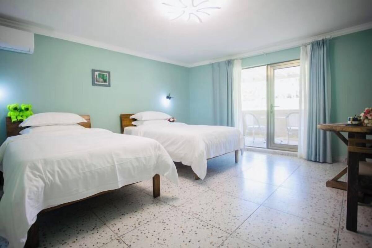 A hotel room with two beds, a dining table, a window with curtains, and a ceiling light fixture.