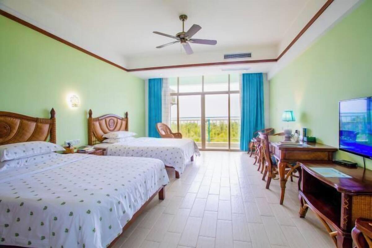 A hotel room with two beds, a ceiling fan, a TV, a desk, and a balcony with blue curtains.