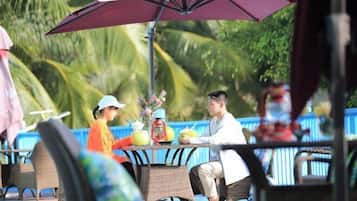 Comedor con sillas de mimbre y una mesa, un paraguas grande y vistas a palmeras.