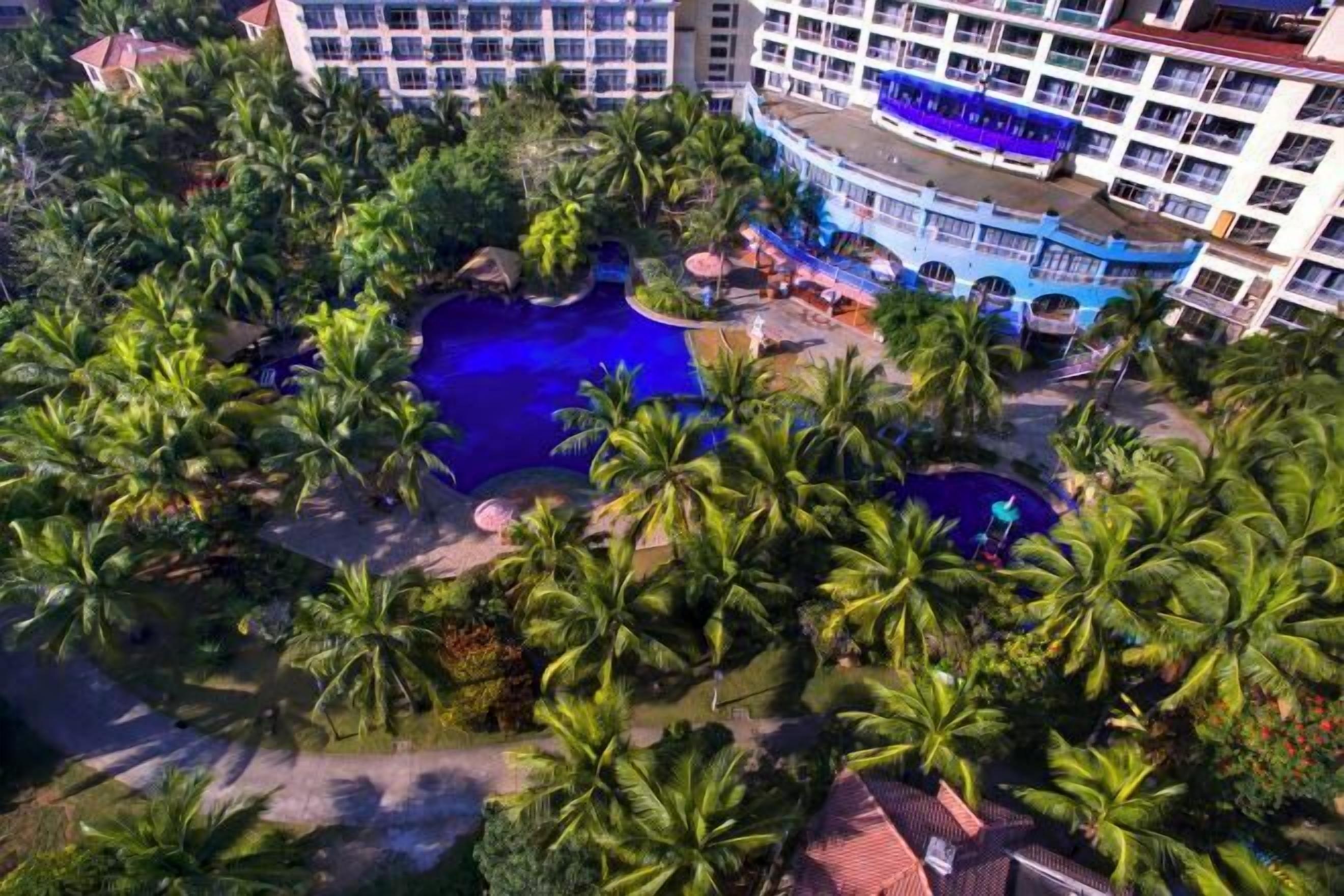 A resort with multiple swimming pools surrounded by palm trees, a blue bus, and a large building with balconies.