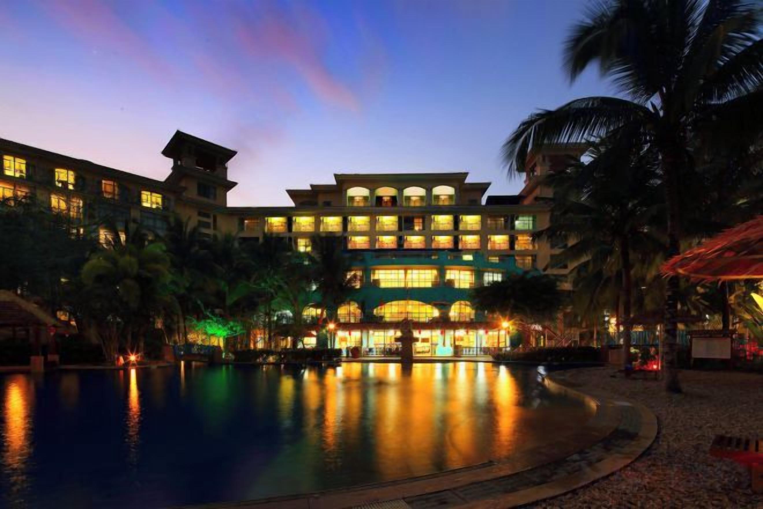 A hotel with a swimming pool, palm trees, and a multi-story building with illuminated windows.