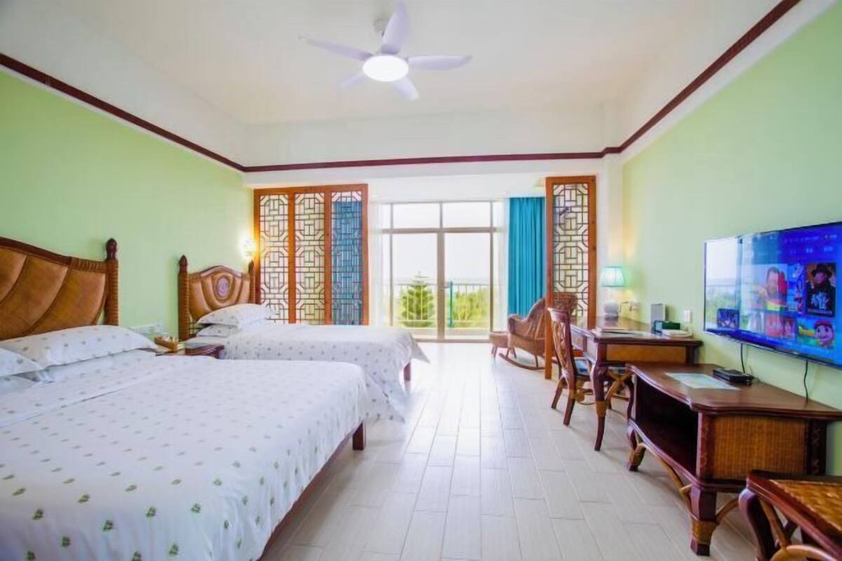 A hotel room with two beds, a desk, a chair, a television, and a ceiling fan.