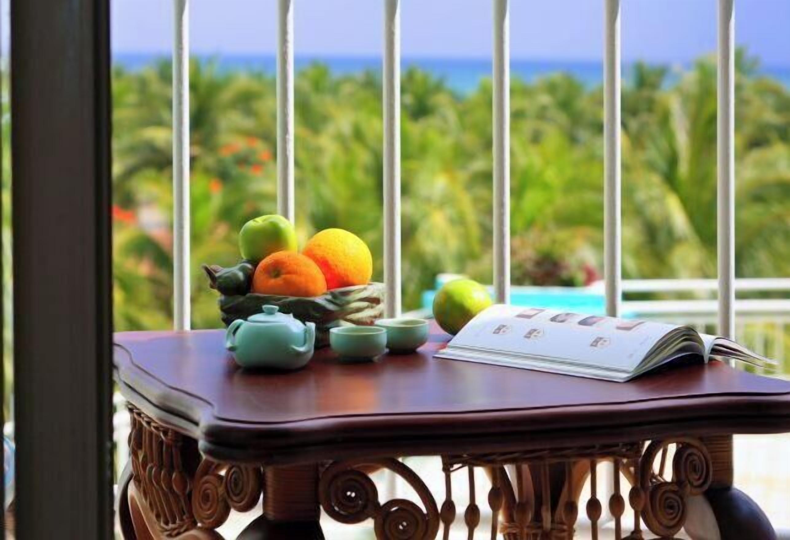 A table with fruits, a teapot, cups, and a magazine on a balcony with a view of palm trees and the ocean.