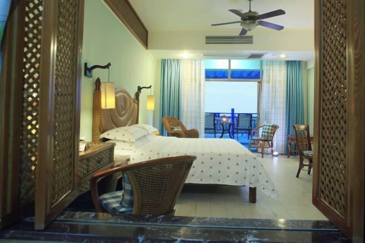 A hotel room with a bed, a chair, a table, a balcony with chairs, and a ceiling fan.