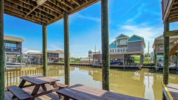 A wooden deck with picnic tables overlooking a calm water body with boats, surrounded by residential buildings. - New For 2025 - Waterfront Bayou Retreat: 25 Min to NOLA. (Barataria)