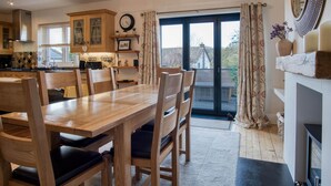 A dining area with a wooden table and chairs, a kitchen in the background, and a view through a glass door. - Little Glen - 3 Bedroom Holiday Home - Tenby (Tenby)