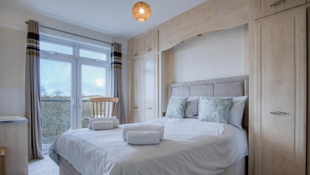 A bedroom with a large bed, a wooden chair, and a view through the window. - Little Glen - 3 Bedroom Holiday Home - Tenby (Tenby)