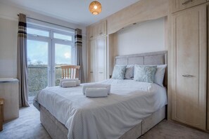 A bedroom with a large bed, a wooden chair, and a view through the window. - Little Glen - 3 Bedroom Holiday Home - Tenby (Tenby)