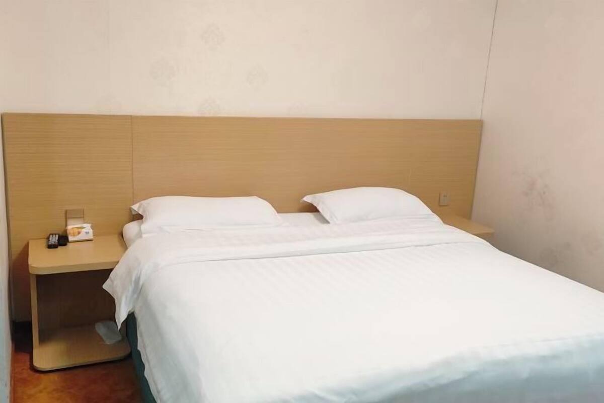 A single bed with a wooden headboard, a bedside table with a lamp and a box, and a plain wall.