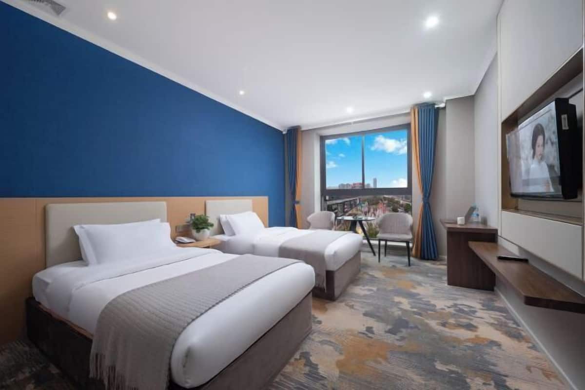 A hotel room with two beds, a TV, a desk, a chair, a window with a city view, and a patterned carpet.