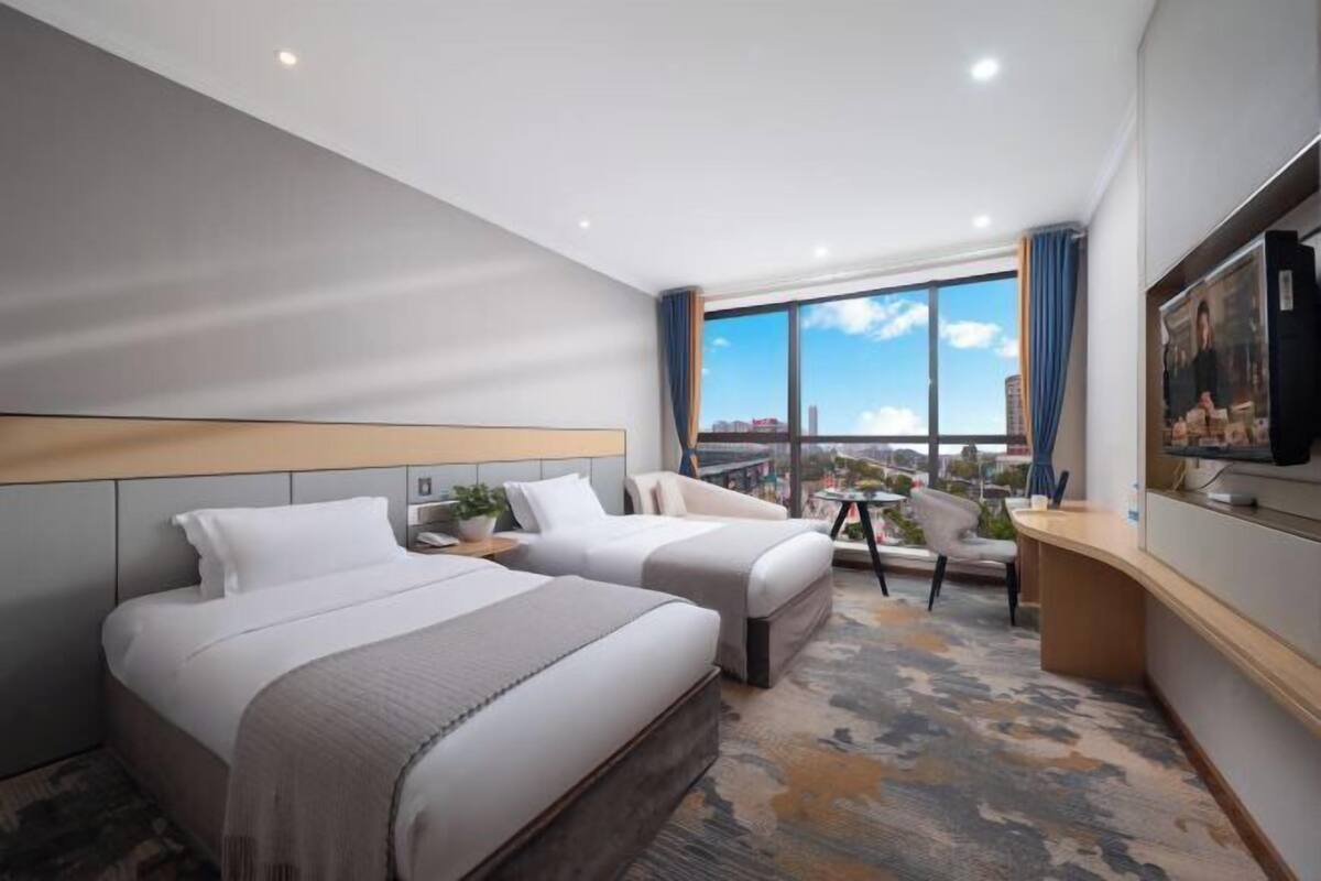 A hotel room with two beds, a desk, a chair, a TV, and a large window with a city view.