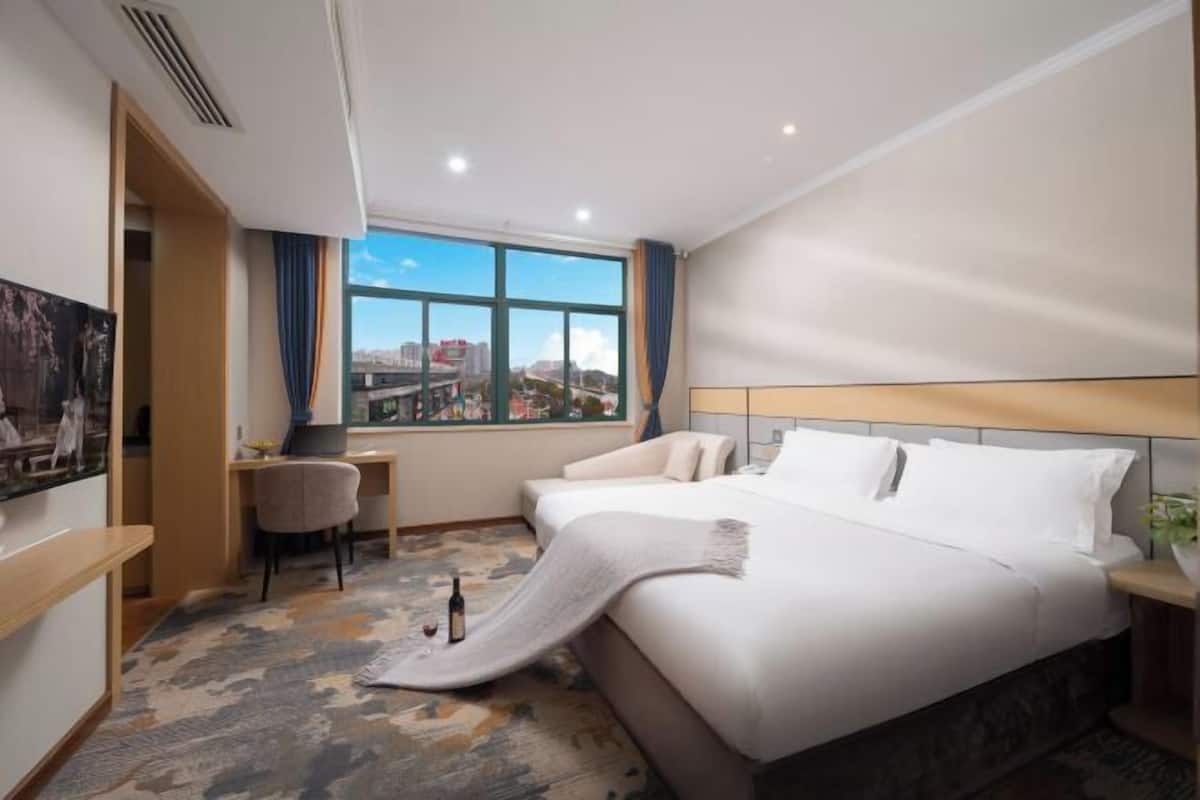 A hotel room with a bed, a desk, a chair, and a large window with a city view.