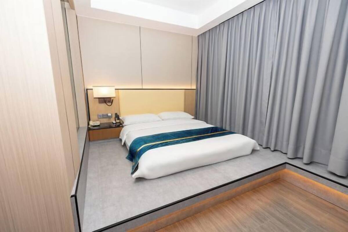 A hotel room with a bed, a nightstand, a lamp, and a view of the sky through a window.