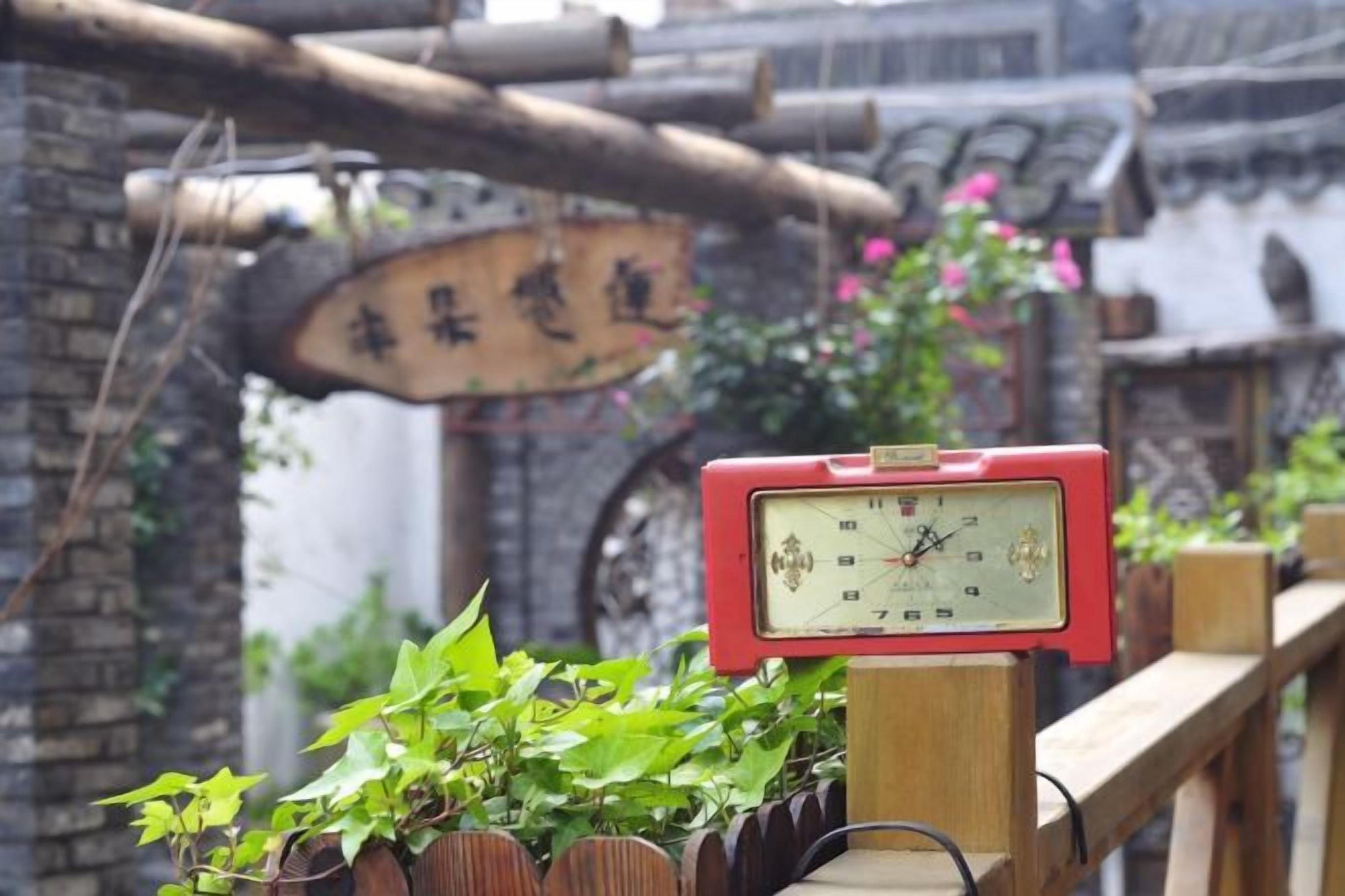 A red clock on a wooden post with a brick wall and a wooden sign in the background.