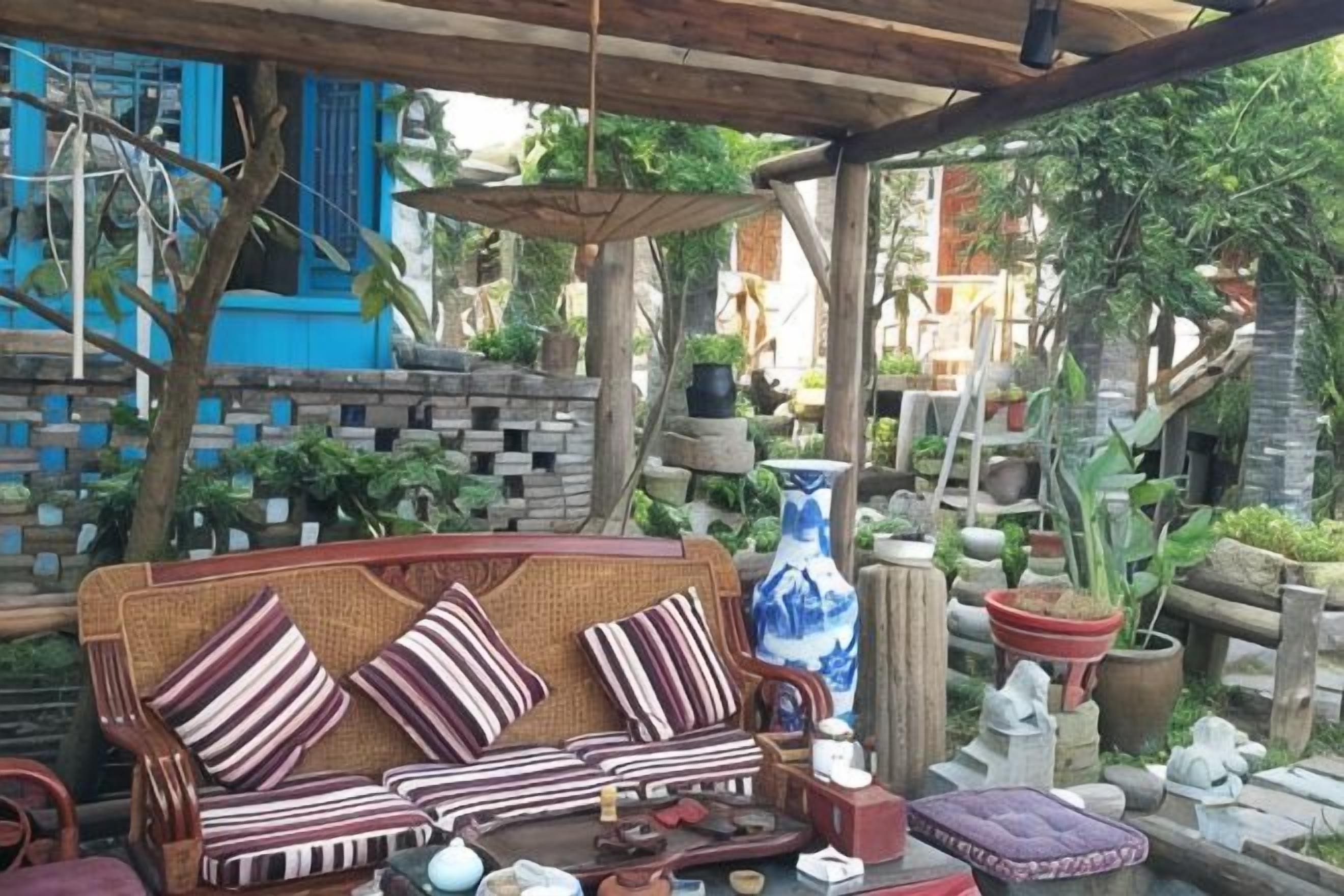 A covered outdoor seating area with a wicker sofa, a large blue vase, and various potted plants.