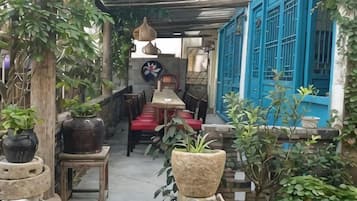 A covered outdoor seating area with tables and chairs, surrounded by potted plants and a blue door.