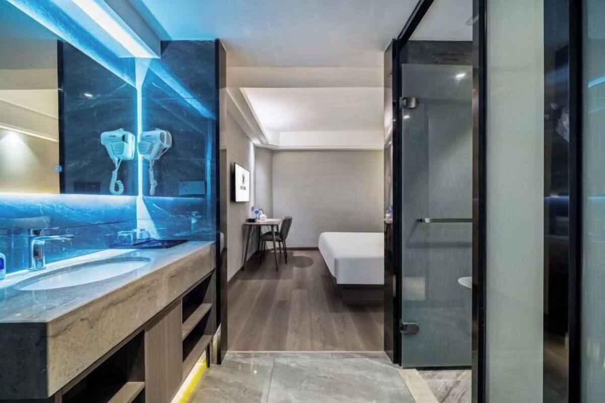 A modern hotel room with a large bathroom vanity, a bed, a small table, and a glass shower enclosure.