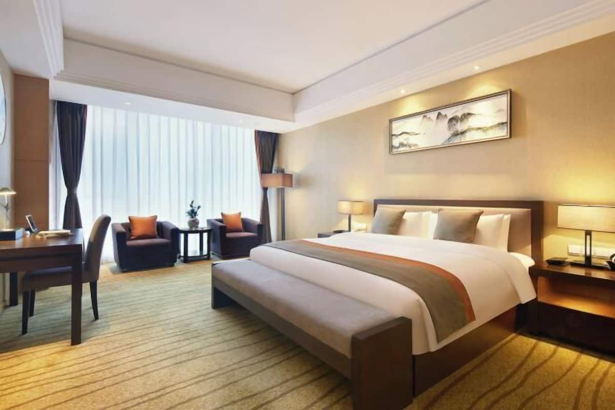 A hotel room with a large bed, a desk, a chair, and a sofa. There is a window with curtains and a painting on the wall.