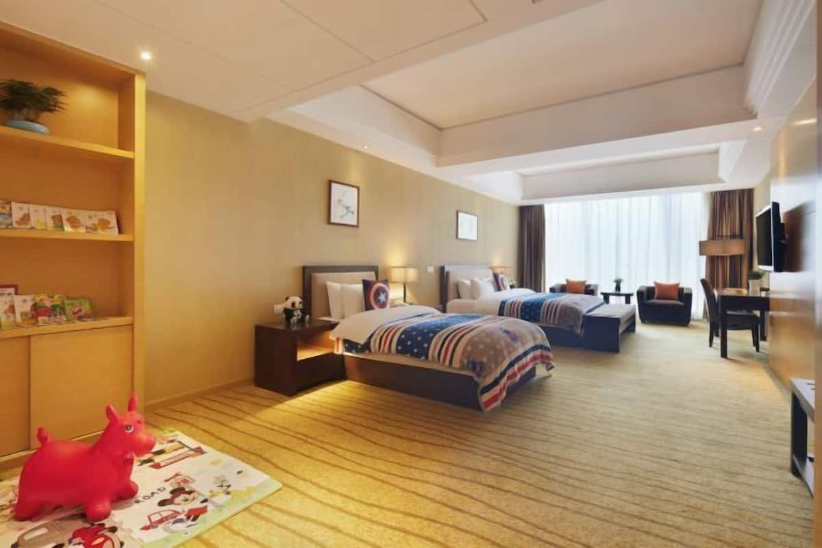 A hotel room with two beds, a desk, a TV, and a bookshelf with children's books.