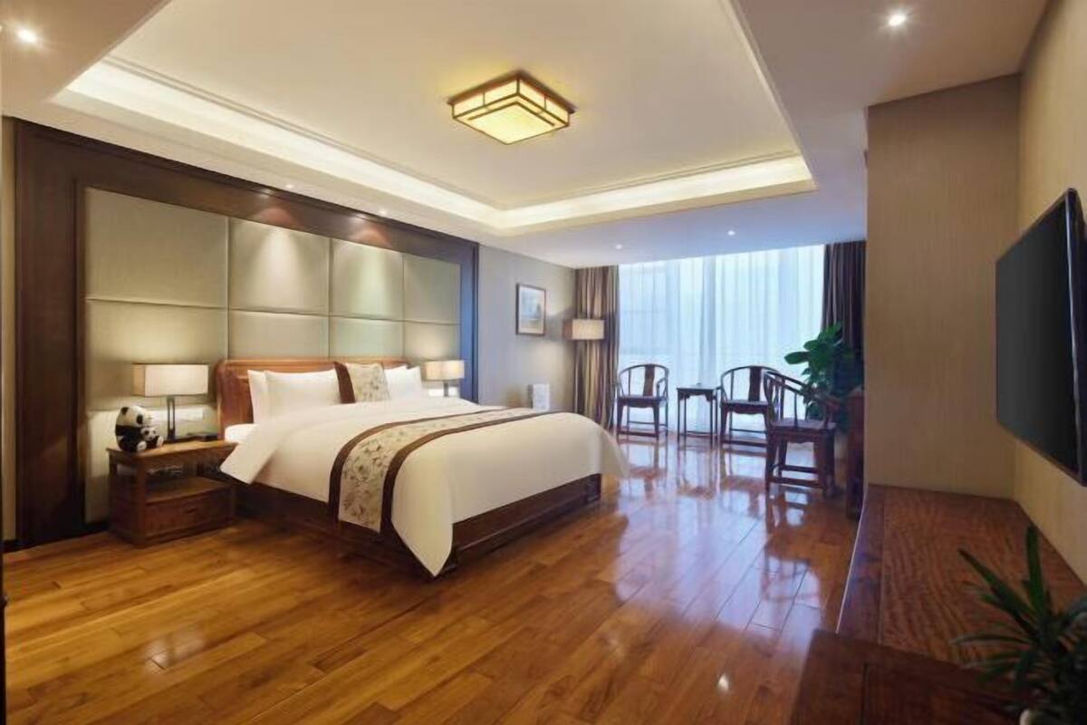 A spacious hotel room with a large bed, a flat-screen TV, a wooden floor, and a balcony with chairs.
