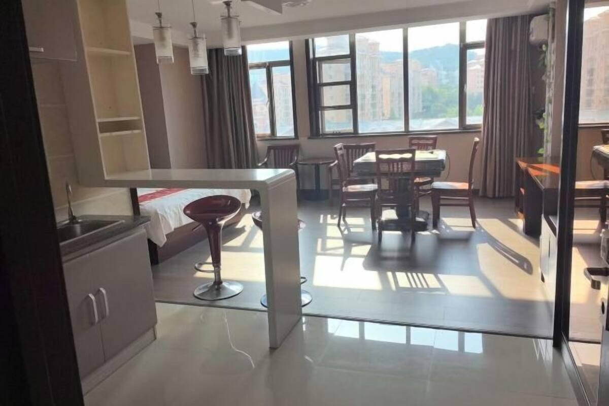 A modern hotel room with a kitchen area, dining space, and large windows offering city views.