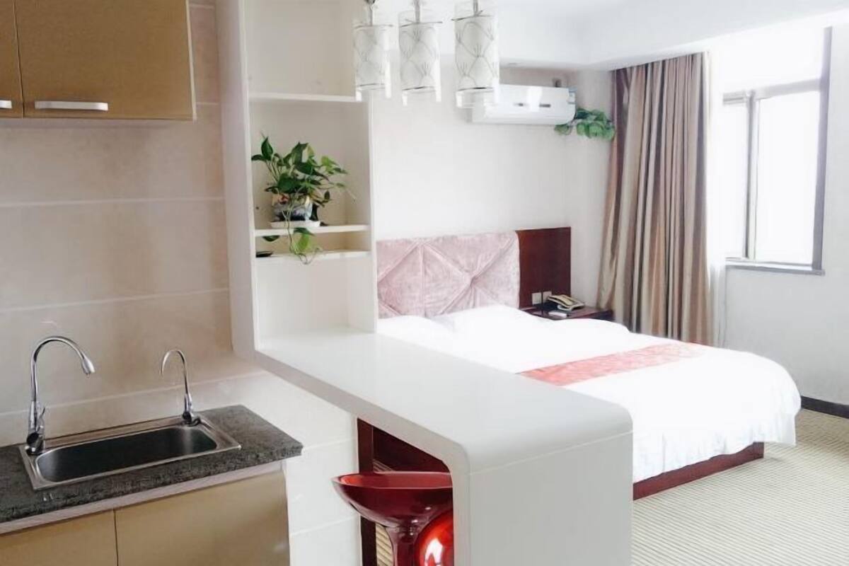 A hotel room with a kitchenette, a bed, a bedside table, a nightstand, a plant, and a window with curtains.