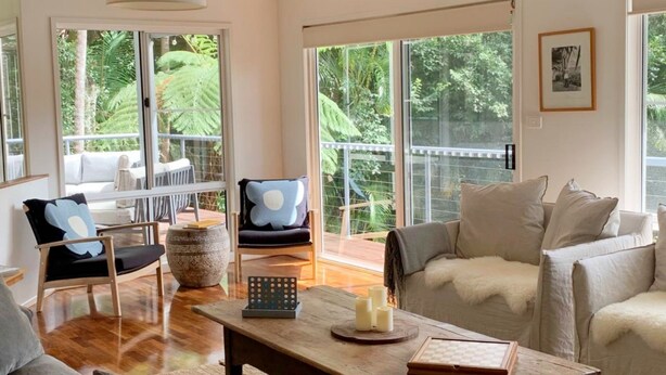 A spacious living room with a wooden coffee table, a sofa, and a chair with a blue pillow. Large windows offer a view of lush greenery outside. - Omanu Shelly Beach (Port Macquarie)