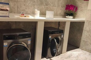A laundry area with two front-loading washing machines under a white counter, a potted plant, and a small sign. - Kunming Southwest Hotel (Kunming)