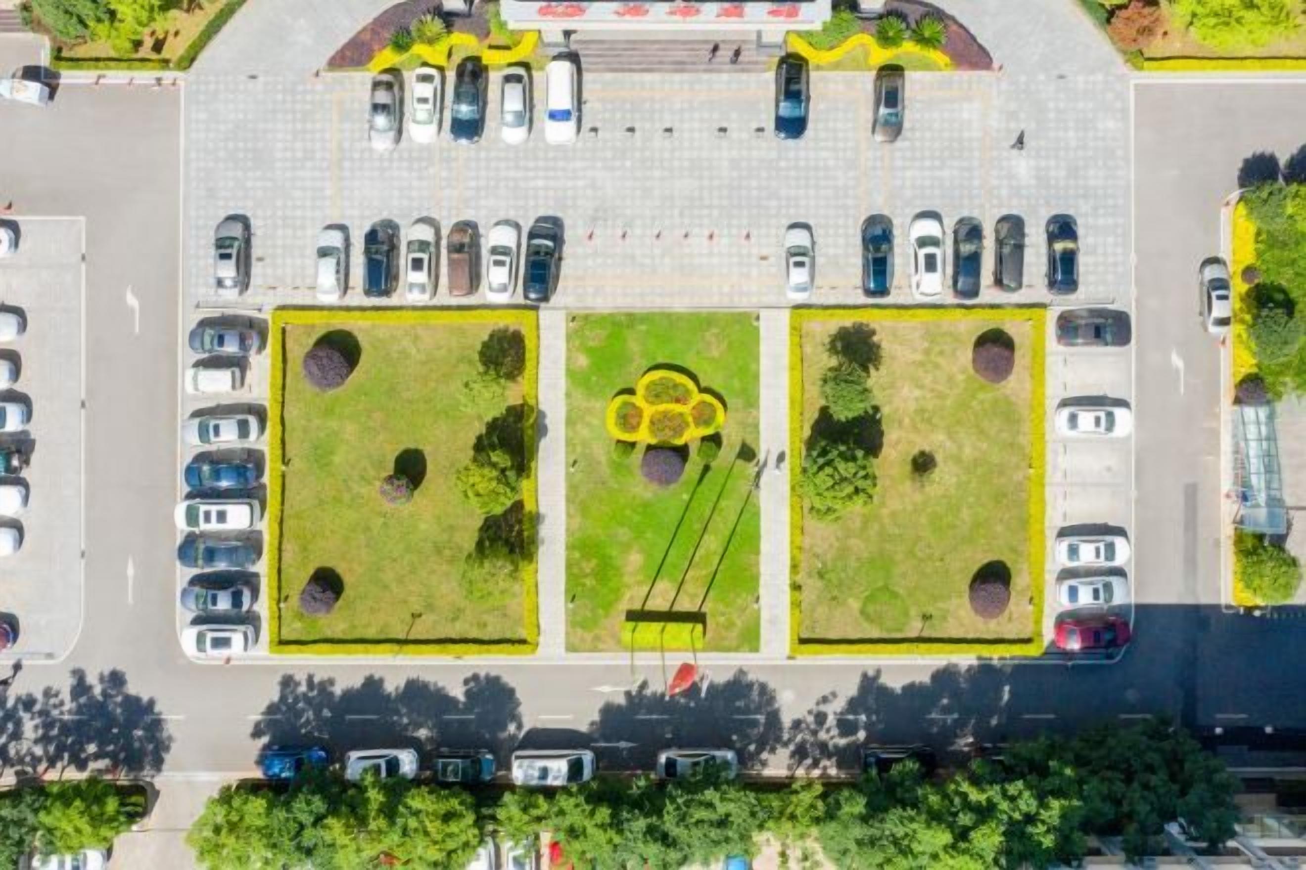 An aerial view of a parking lot with multiple rows of cars, green landscaped areas, and a central circular feature.