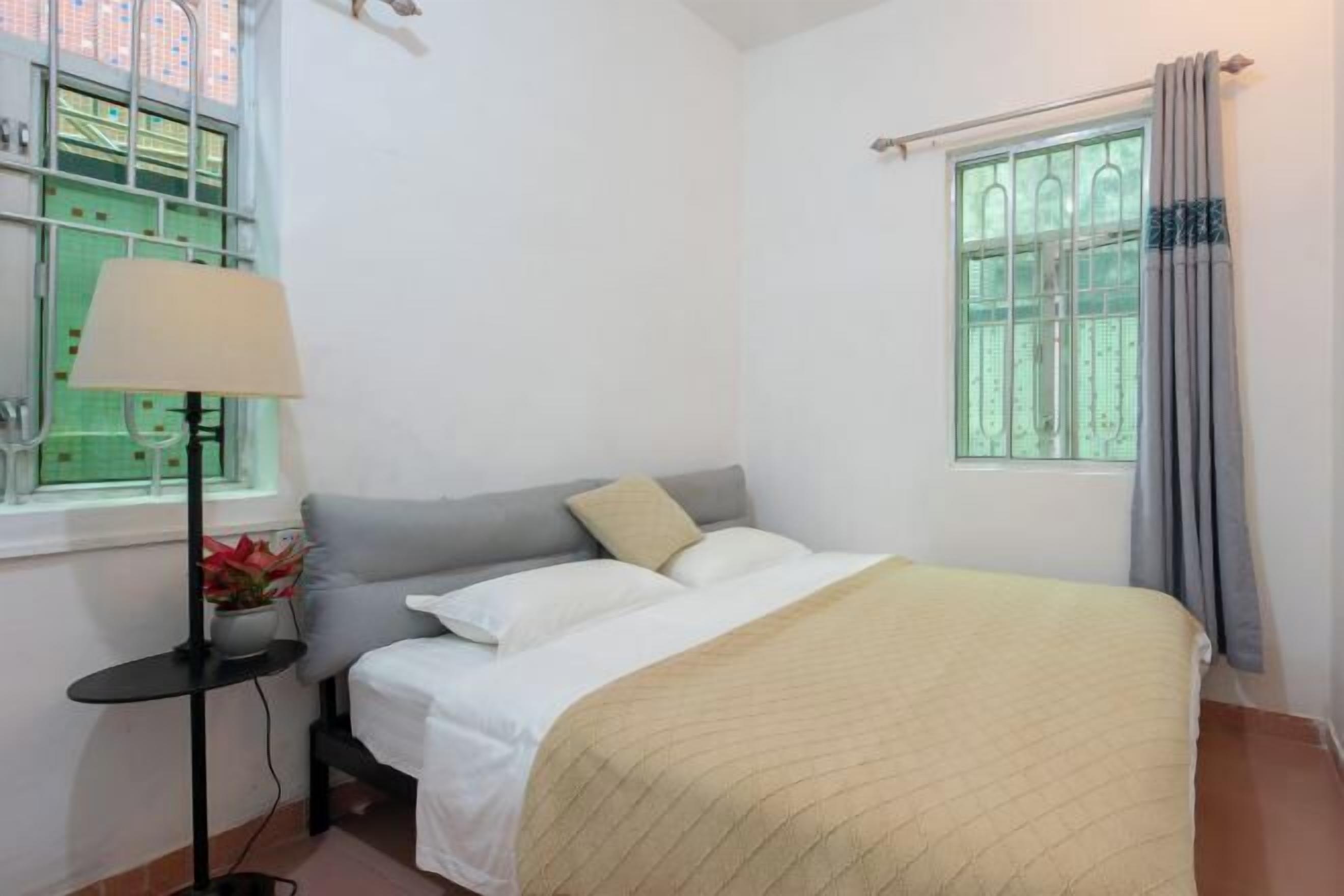 A bedroom with a bed, a bedside table with a lamp, a window with curtains, and a view of a building outside.