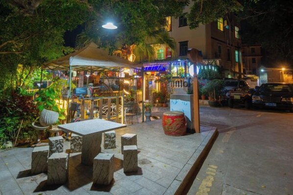 An outdoor seating area with stone tables and chairs, a covered pavilion, and a street view with parked cars and buildings in the background. - HaiBei Hostel (Shenzhen)