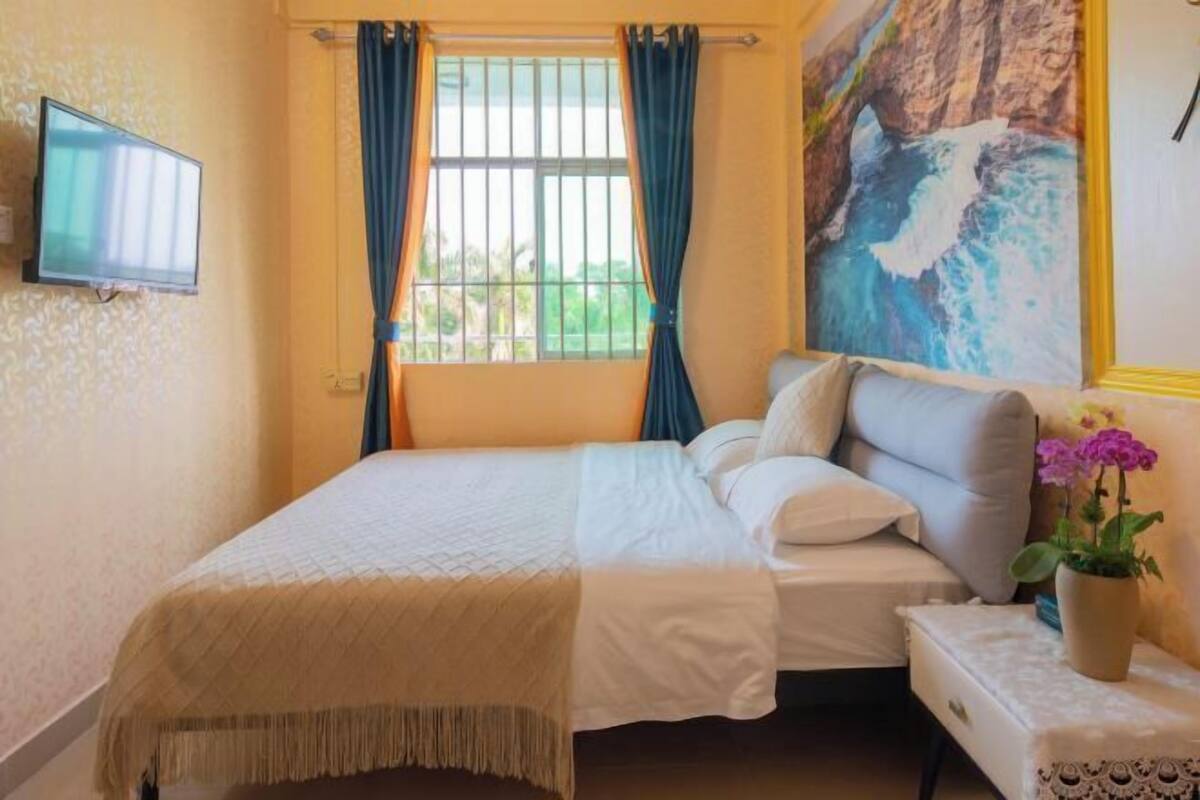 A bedroom with a bed, a TV mounted on the wall, a window with curtains, and a painting of a coastal scene.