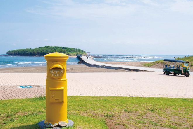 Una playa con un poste amarillo, un camino pavimentado y un vehículo a lo lejos.