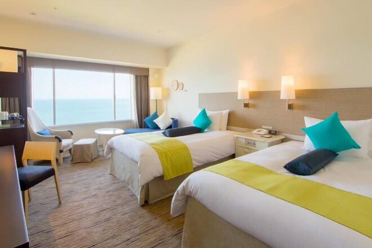 A hotel room with two beds, a desk, a chair, a small table, and a view of the sea.