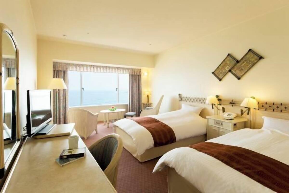 A hotel room with two beds, a desk, a chair, a TV, and a window with a view.