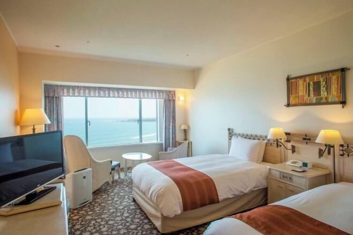 A hotel room with a bed, bedside tables, a desk with a computer, a chair, a small table, and a window with curtains.