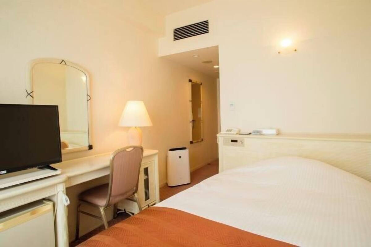 A hotel room with a bed, a desk with a chair, a television, a lamp, a mirror, and a small bedside table with a phone and a remote control.