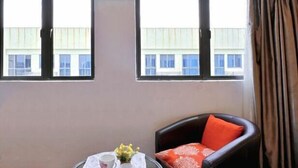 Une pièce avec deux fenêtres, une chaise avec un coussin rouge et une petite table recouverte d’une nappe blanche, sur laquelle se trouve un vase de fleurs.